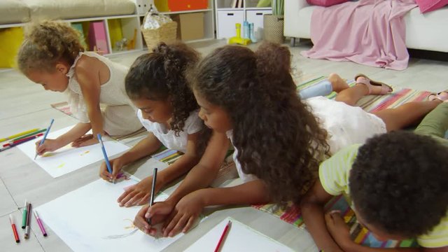 Four black children of elementary school age lying on floor at home and using colored pencils to draw pictures