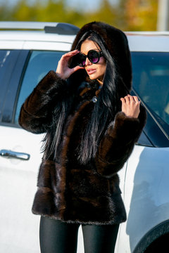 Beautiful sexy brunette in sunglasses and a fur coat walks down the street on sunny day and looks away. Late autumn or early winter. Vertical fashionable portrait