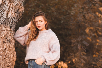 Smiling teenage girl 17-18 year old wearing knitted sweater posing outdoors. Looking at camera. Autumn season. 20s.