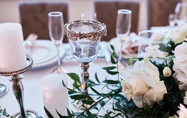 Wedding table serivce. Candles on silver vases and glasses with water stand among white flowers on wedding dinner table