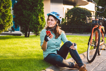 Man and city rolling bicycle, environmentally friendly transport. Beautiful young caucasian woman worker sitting resting on the grass uses a red mobile phone near an orange bicycle with a coryne.