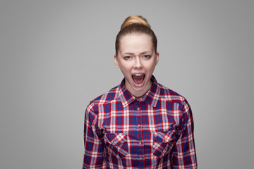 Angry beautiful blonde girl in red, pink checkered shirt, collected bun hairstyle, makeup standing and looking at camera with and screaming. indoor studio shot. isolated on gray background