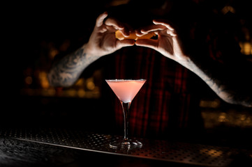 Barman sprinkling orange peel juice into a Cosmopolitan cocktail in dark background