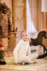Christmas portrait of happy blonde child girl is sitting at home, decorated tree, presents and lights on background. New Year and Christmas Holidays