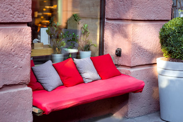 a bench with a red soft mattress and pillows of red and gray at the window of the facade of the building, a place to rest on a city street.