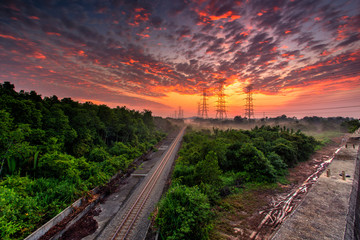 Railroad against beautiful burning sky at sunrise. Industrial landscape with railroad, colorful blue sky with burning clouds, sun, trees and green grass. Railway junction.