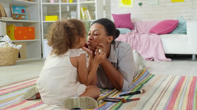 Cute little black girl of primary school age sitting on floor and applying lipstick on mother’s lips, mother kissing her