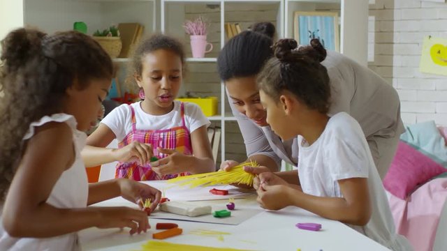Three cute black girls sitting at desk in art studio and creating objects with modeling clay, their female teacher bringing spaghetti to use in crafts