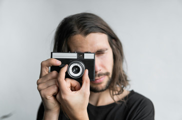 Handsome young bearded man with a long hair and in a black shirt holding vintage old-fashioned film camera on a white background and looking in camera viewfinder.