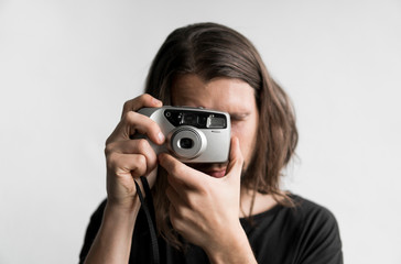 Handsome young bearded man with a long hair and in a black shirt holding vintage old-fashioned film camera on a white background and looking in camera viewfinder.