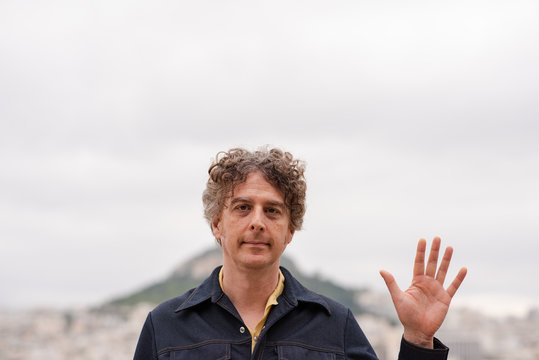 Portrait Of A Mature Man Waving To The Camera Saying Hello Or Welcome, After Reaching The Top Of A Mountain.