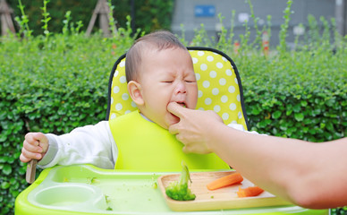 Close-up infant baby boy sitting on kid chair eating with something stuck in his mouth and mother...