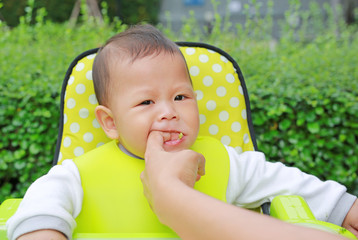 Close-up infant baby boy sitting on kid chair eating with something stuck in his mouth and mother help to keep out.
