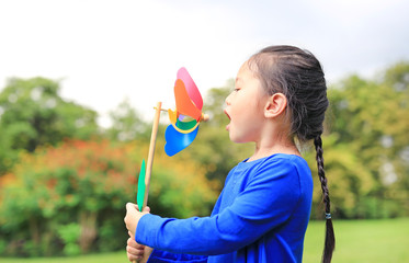 Portrait of little Asian kid girl blowing wind turbine in the summer garden.
