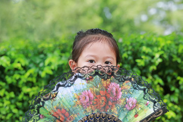 Portrait of little Asian child girl holding chinese style fan hide her face in the summer garden. Close-up.