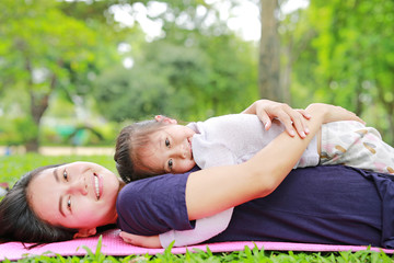 Happy Asian mom embrace her daughter lying in the green garden with looking camera. Funny mother and child girl playing in summer park.