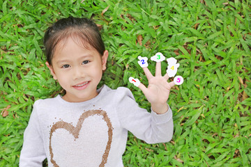 Smiling little Asian child girl lying on green grass lawn with showing white stickers with number one to five on her fingers.