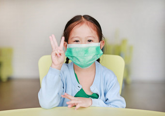 Cute little Asian child girl wearing a protective mask with showing three fingers sitting on kid chair in children room.