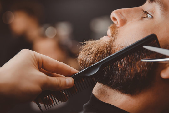 Hipster man sitting in armchair barber shop while hairdresser shaves beard with scissors.