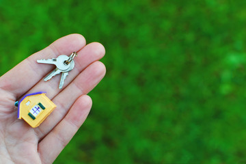 Hand holding or giving small colorful house model and keys on green blurred grass background with copy space for text. Real estate, buying moving new home or renting property or apartments concept.