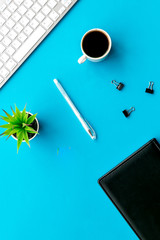 Office desk of creative person or hipster.Cute workplace. Keyboard and glasses near coffee, notebook, green room plant and stationery on blue, turquoise background top view copy space