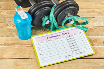 Healthy lifestyle concept. Mock up on workout and fitness dieting diary with copy space. Slimming diary sheet, measuring tape, blue shaker and dumbbells on a rustic wooden table
