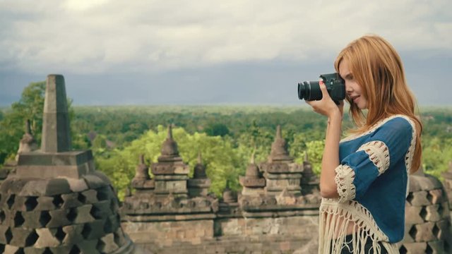 Young caucasian happy woman taking photo with camera on top of Borobudur temple in front of stone stupas and view to forest in Java, Indonesia