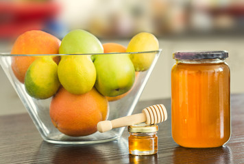 Big and small glass jar of honey with wooden sipper spoon and bowl of fruits on the background.