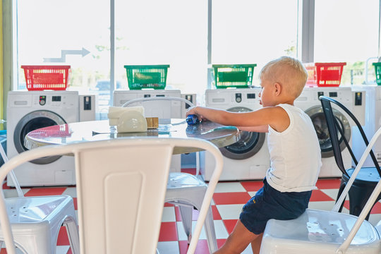 A Little Boy Sitting At A Table And Waiting For The End Of Washing In The Laundromat. Room With Laundry Machines, Sunny Summer Day.