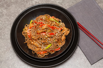 Fried noodles with vegetables, chicken and sesame in a black bowl. Gray background. Close up