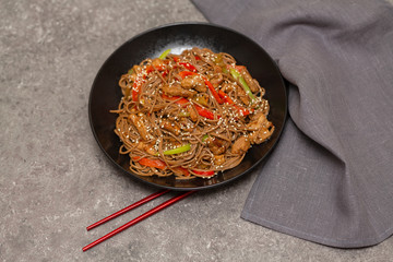 Fried noodles with vegetables, chicken and sesame in a black bowl. Gray background. Close up