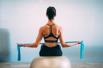 Fotobehang Young sporty attractive woman practicing pilates with resistance bands back sitting on fitness balls on white urban studio. Yoga and Pilates lifestyle healthy concept. © ManuPadilla