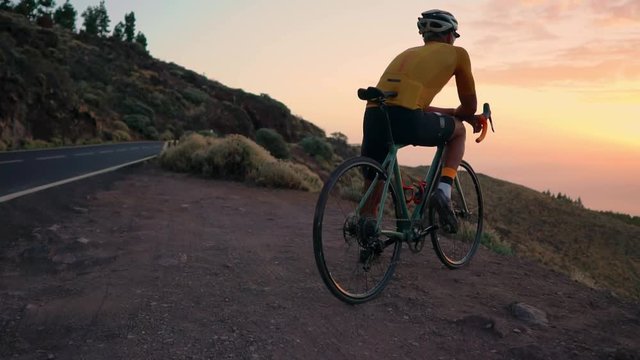 Young sports man sitting on a bike on a mountain top in a yellow t-shirt helmet and sports equipment looking at the beautiful view of the mountains and the setting sun. Rest after training . Slow
