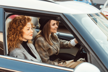 side view of happy stylish woman in black hat driving car while her ginger female friend sitting near