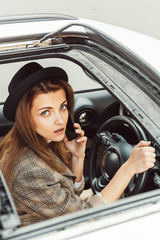 portrait of stylish woman in black hat talking on smartphone while sitting at steering wheel in car