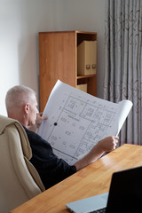 Naklejka premium Side view of professional mature male engineer sitting in office chair and taking a good look at blueprint of painted ceiling