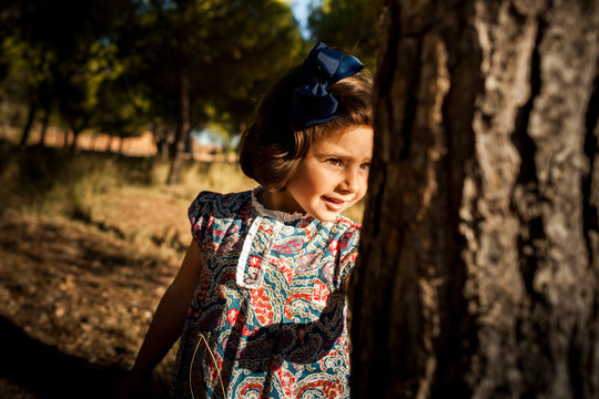 ni&ntilde;a asomada detr&aacute;s de un tronco en mitad de un pinar  ba&ntilde;ada por el sol del atardecer