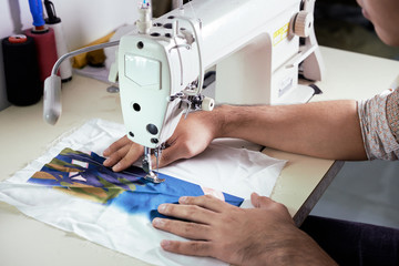 Close-up of male designer sitting at the table and using sewing machine to make clothing