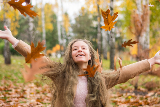 A teenager girl in a coat laughs and throws up leaves in autumn park against the background of yellow foliage.