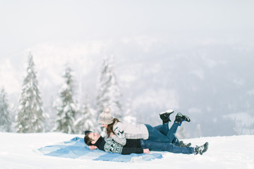 Charming young couple in warm winter knitted sweaters lying on the wool blue chekered blanket in snowy forest. Love in winter, Happy New Year and Christmas concept