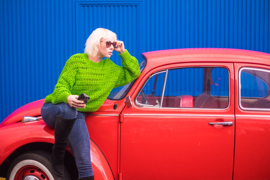 Fashion coloured concept image with blonde caucasian girl standing near a red vintage old car with blue steel wall in backgorund. Urban and colors with people travel technology concept