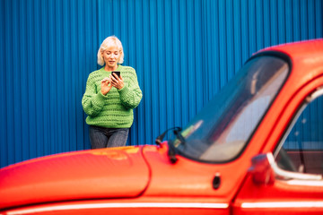 Colors and alternative happy lifestyle concept image with blonde attractive woman thirty years old using mobile phone internet technology outside the red car and with blue wall on the background