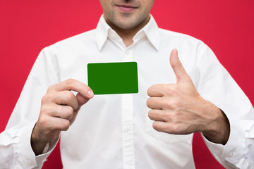 Attractive businessman  in white shirt showing business card to the camera, shows thumb up, red background, with copy space, for advertisement, front view, close up