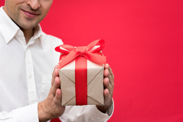 Attractive man in white shirt holding a box with a gift with a red ribbon, on camera, bright red background, with copy space, for advertisement, front view, close up