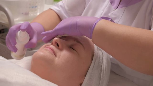 A professional cosmetologist in lilac gloves sprays a moisturizing tonic on the face of a European woman of middle age. Cosmetic procedure in a beauty salon. High quality raw video.