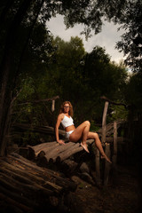 A beautiful slim girl with red hair in a white T-shirt and white shorts sits on the bridge of logs in a dark forest.