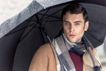 handsome man in coat and scarf with umbrella looking at camera on street