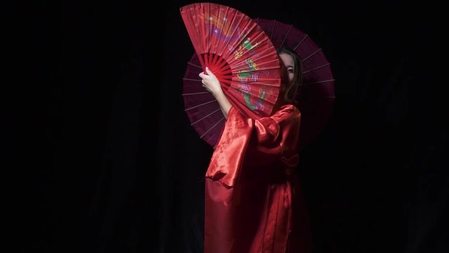 Lovely geisha appears from the dark, holding a fan and an umbrella