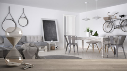 Wooden table, desk or shelf with crystal modern hourglass measuring the passing time and house keys over modern scandinavian living room, architecture interior design, copy space background