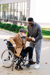 Obraz premium young african american nurse helping senior disabled man to get up from wheelchair on street